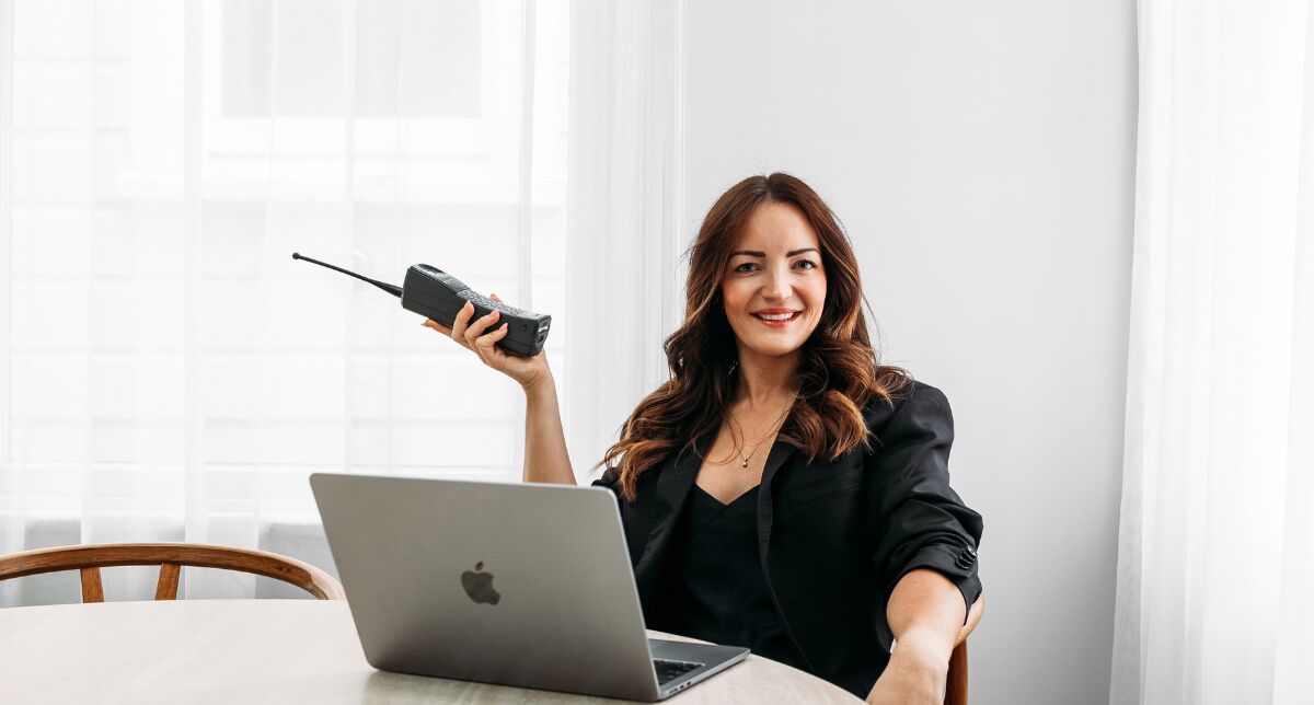 Christine Westbury smiling at a laptop, holding up a retro mobile phone, ready for a sales conversation